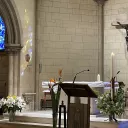 Titre Messe sur fond rose de rubrique Vie spirituelle accompagné du titre lieu à l'espace Bernadette avec sur la droite de la vignette une croix chrétienne blanche et au pied un ensemble d'objets utiles à la communion durant les célébrations eucharistiques