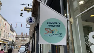 Drapeau de l'association des commerçants "shop'in Alençon" flottant sur la devanture d'un magasin d'habillement.